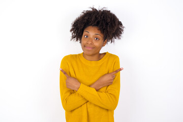 young beautiful African American woman wearing yellow sweater against white wall crosses arms and points at different sides hesitates between two items or variants. Needs help with decision