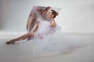 Naklejka premium Romantic beauty woman in a light transparent dress is reflected in a flexible mirror sitting on the floor. Perfect body and long legs