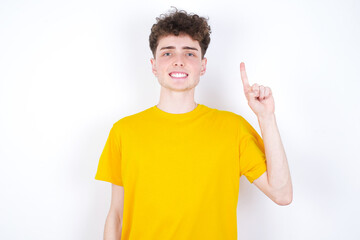 young caucasian handsome man with curly hair wearing yellow T-shirt against white studio background  showing and pointing up with finger number one while smiling confident and happy.