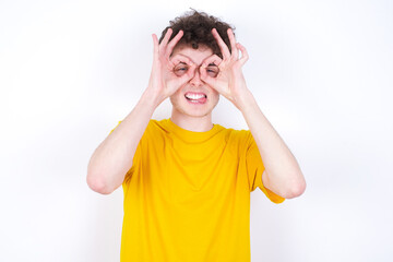 young caucasian handsome man with curly hair wearing yellow T-shirt against white studio wall doing ok gesture like binoculars sticking tongue out, eyes looking through fingers. Crazy expression.