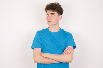 Fototapeta premium Charming thoughtful young caucasian handsome man with curly hair wearing blue T-shirt against white studio wall stands with arms folded concentrated somewhere with pensive expression thinks what to do