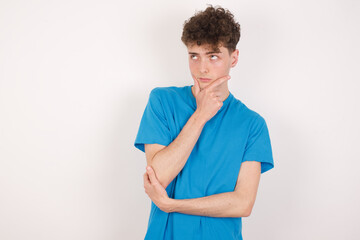 Thoughtful young caucasian handsome man with curly hair wearing blue T-shirt against white studio background holds chin and looks away pensively makes up great plan