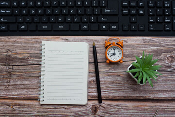 Notepad with pen, alarm clock, potted plan and keyboard on wooden desk