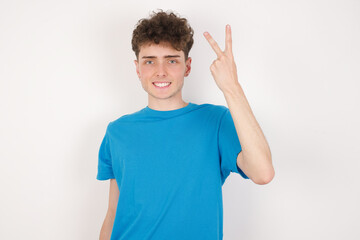 Fototapeta premium young caucasian handsome man with curly hair wearing blue T-shirt against white studio background smiling and looking friendly, showing number two or second with hand forward, counting down