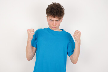 Fototapeta premium Irritated young caucasian handsome man with curly hair wearing blue T-shirt against white studio blows cheeks with anger and raises clenched fists expresses rage and aggressive emotions. Furious model