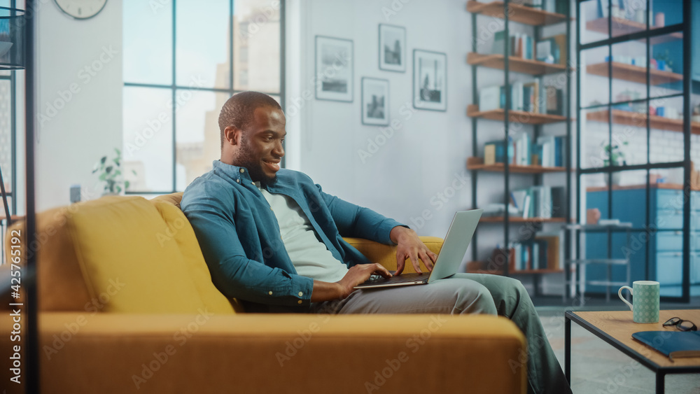 Poster handsome black african american man working on laptop computer while sitting on a sofa in cozy livin