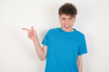 Fototapeta premium young caucasian handsome man with curly hair wearing blue T-shirt against white studio background points aside on copy blank space. People promotion and advertising concept