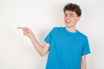young caucasian handsome man with curly hair wearing blue T-shirt against white laughs happily points away on blank space demonstrates shopping discount offer, excited by good news or unexpected sale.