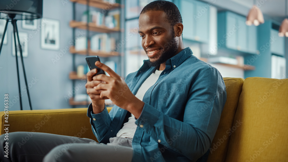 Poster handsome black african american man using smartphone while sitting on a sofa in cozy living room. fr