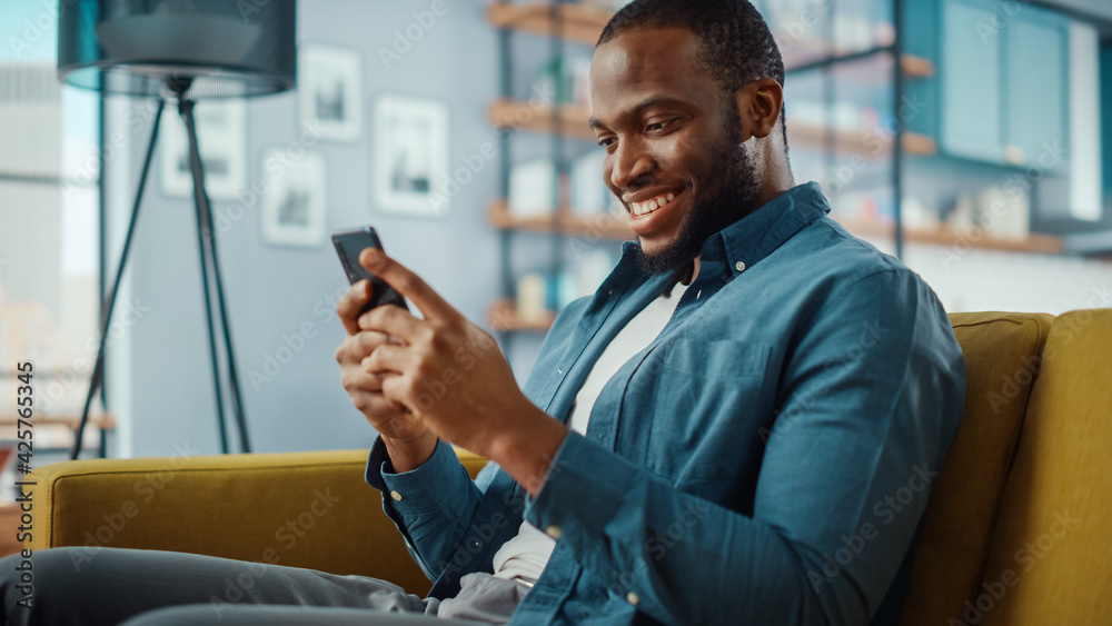 Poster handsome black african american man using smartphone while sitting on a sofa in cozy living room. fr