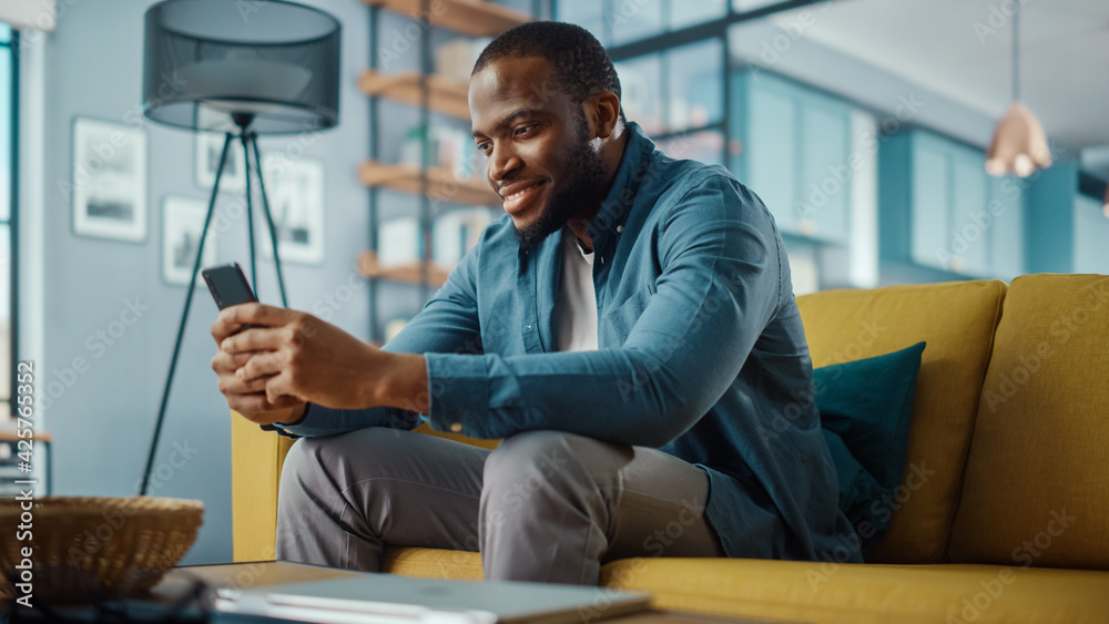 Poster handsome black african american man using smartphone while sitting on a sofa in cozy living room. fr