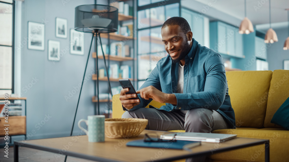 Poster handsome black african american man using smartphone while sitting on a sofa in cozy living room. fr