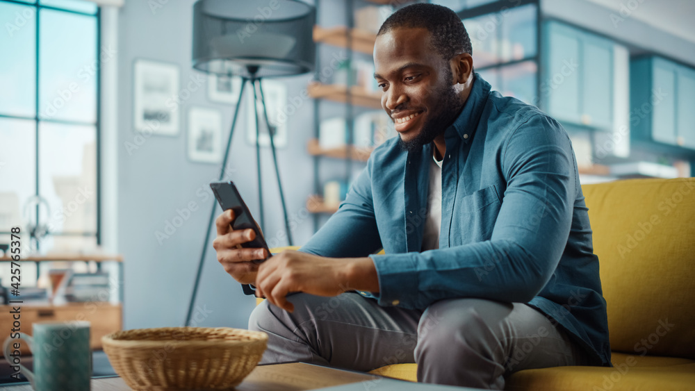 Poster handsome black african american man using smartphone while sitting on a sofa in cozy living room. fr