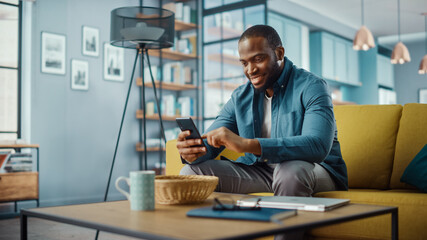 Fototapeta na wymiar Handsome Black African American Man Using Smartphone while Sitting on a Sofa in Cozy Living Room. Freelancer Working From Home. Browsing Internet, Using Social Networks, Having Fun in Flat.
