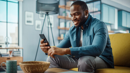 Handsome Black African American Man Using Smartphone while Sitting on a Sofa in Cozy Living Room....