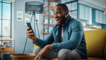 Excited Black African American Man Having a Video Call on Smartphone while Sitting on a Sofa in...