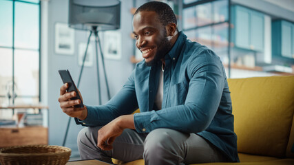 Excited Black African American Man Having a Video Call on Smartphone while Sitting on a Sofa in Living Room. Happy Man Smiling at Home and Talking to Colleagues and Clients Over the Internet. - obrazy, fototapety, plakaty
