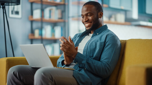 Handsome Black African American Man Working From Home And Having Video Call On Laptop Computer While Sitting On A Sofa In Living Room. He Is Clapping His Hands In Celebration Of The Company's Success.