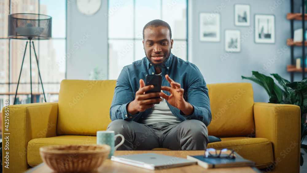 Aufkleber Excited Black African American Man Using Smartphone while Resting on a Sofa in Living Room. Happy Man Smiling at Home and Chatting to Colleagues and Clients Over the Internet. Using Social Networks.