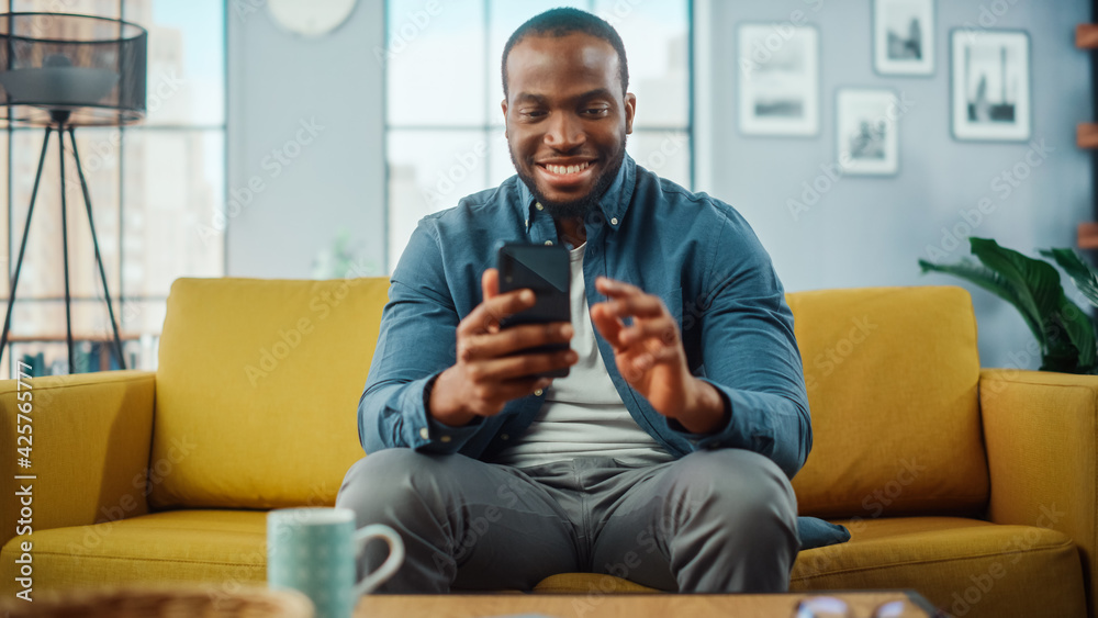Poster excited black african american man using smartphone while resting on a sofa in living room. happy ma
