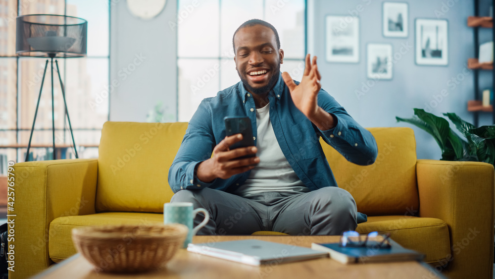 Poster happy black african american man having a video call on smartphone while sitting on a sofa in living