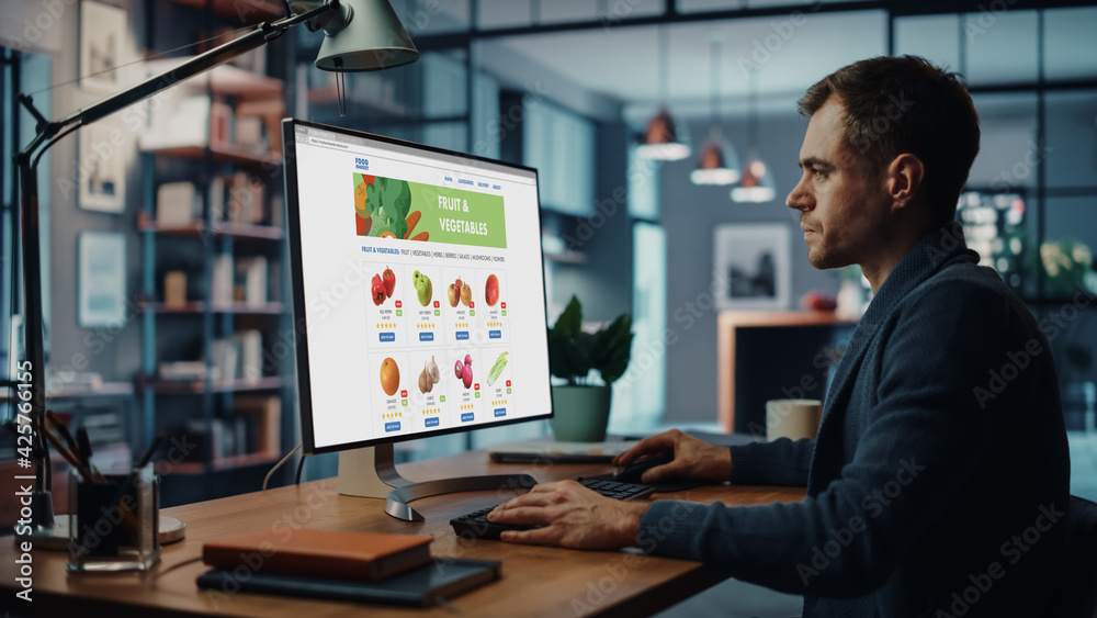 Poster handsome caucasian male is using desktop computer with groceries delivery web page, choosing fresh v