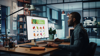 Handsome Black African American is Using Desktop Computer with Groceries Delivery Web Page to Order Fresh Vegetables to Make Dinner. He Lives in Modern Huge Appatment.