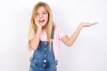 Crazy beautiful caucasian little girl wearing jeans overall over white background advising discount prices hold open palm new product