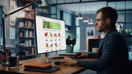Handsome Caucasian Male is Using Desktop Computer with Groceries Delivery Web Page, Choosing Fresh Vegetables for Order to Make Dinner. He Lives in Huge Modern Apartment.