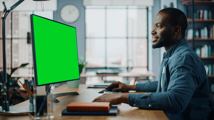 Handsome Black African American Specialist Working on Desktop Computer with Green Screen Mock Up Display at Home Living Room. Freelance Man Chatting to Clients Over the Internet on Social Networks.