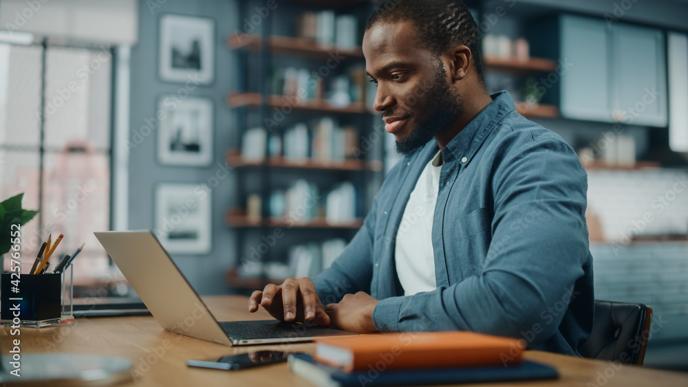 Poster handsome black african american man working on laptop computer while sitting behind desk in cozy liv