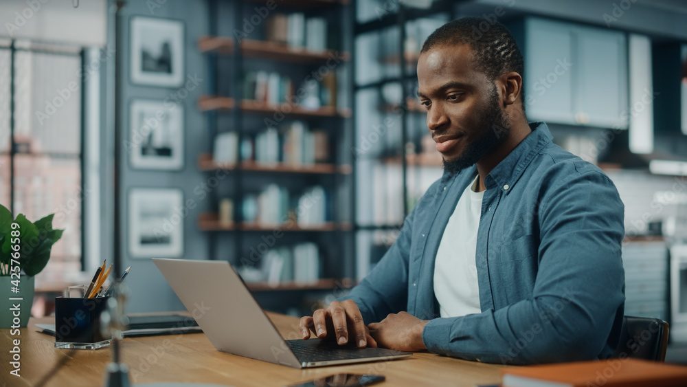 Poster handsome black african american man working on laptop computer while sitting behind desk in cozy liv