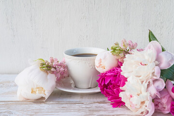 White cup of coffee and bunch of pink and white peonies on white paint wooden background. Space for text