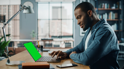 Fototapeta na wymiar Handsome Black African American Specialist Working on Laptop Computer with Green Screen Mock Up Display at Home Living Room. Freelance Man Chatting to Clients Over the Internet on Social Networks.