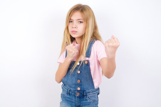 beautiful caucasian little girl wearing denim jeans overall over white background Ready to fight with fist defense gesture, angry and upset face, afraid of problem.