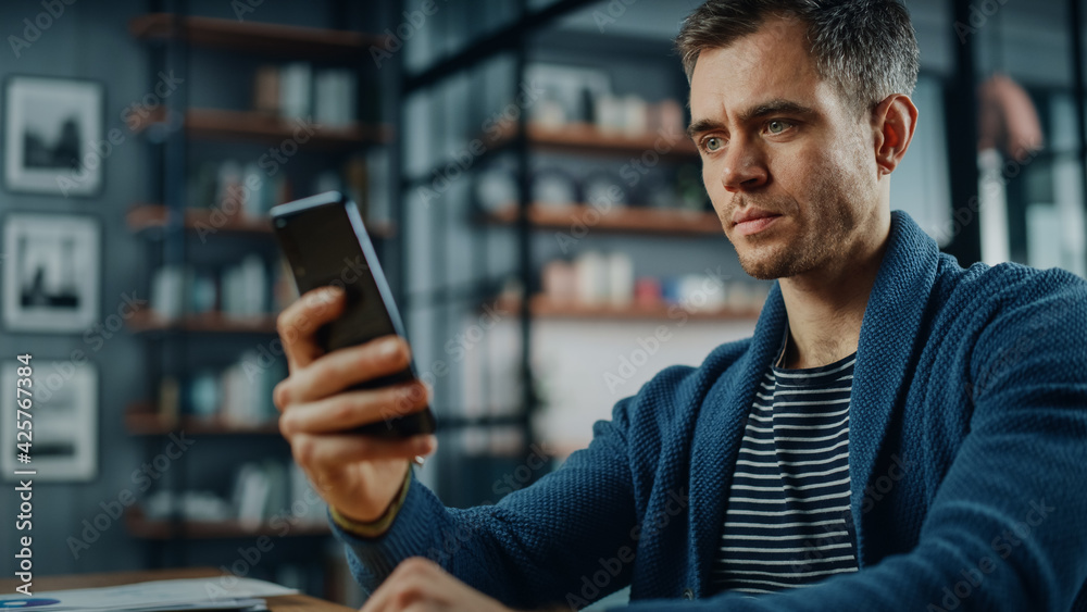 Aufkleber Serious Caucasian Man Using Smartphone while Sitting at a Desk in Stylish Living Room. Young Man Smiling at Home and Chatting to Colleagues and Clients Over the Internet. Using Social Networks.