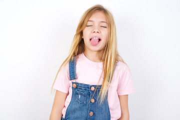 beautiful caucasian little girl wearing denim jeans overall over white background sticking tongue...
