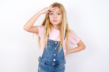 Fototapeta premium beautiful caucasian little girl wearing denim jeans overall over white background having problems, worried and stressed holds hand on forehead.