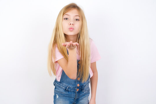 beautiful caucasian little girl wearing denim jeans overall over white background looking at the camera blowing a kiss with hand on air being lovely and sexy. Love expression.