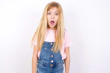 Oh my God. Surprised beautiful caucasian little girl wearing denim jeans overall over white background stares at camera with shocked expression exclaims with unexpectedness,