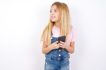 beautiful caucasian little girl wearing denim jeans overall over white background holding a smartphone and looking sideways at blank copyspace.