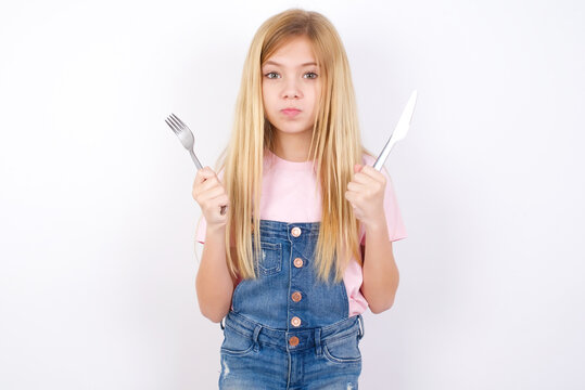 Model hungry beautiful caucasian little girl wearing denim jeans overall over white background holding in hand fork knife want tasty yummy pizza pie