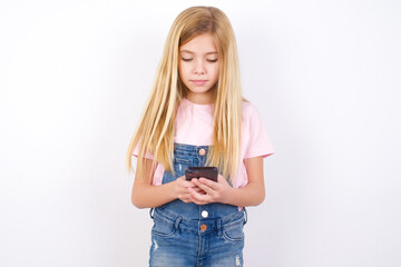 Excited beautiful caucasian little girl wearing denim jeans overall over white background winking and eye hold smart phone use read social network news