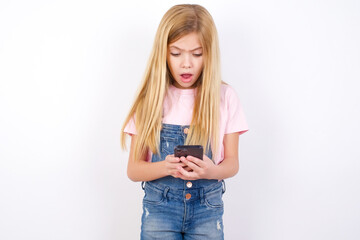 Focused beautiful caucasian little girl wearing denim jeans overall over white background use smartphone reading social media news, or important e-mail