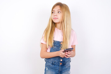 beautiful caucasian little girl wearing denim jeans overall over white background holds telephone hands reads good youth news look empty space advert