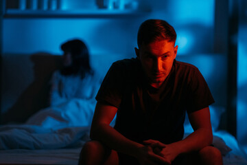 Couple conflict. Domestic abuse. Bad relationship. Aggressive man sitting on bed with afraid insecure woman victim in dark night red blue neon light on defocused background.