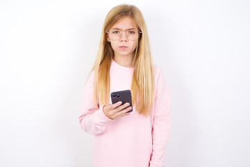 Naklejka premium Upset dissatisfied beautiful caucasian little girl wearing pink sweater uses mobile software application and surrfs information in internet, holds modern mobile hand