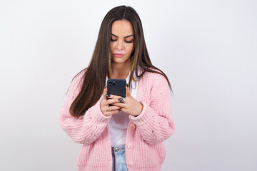 Excited young beautiful caucasian woman wearing pink knitted jacket over white wall winking and eye...