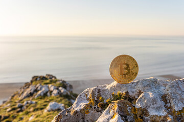 Close-up of Bitcoin on a stone outdoor with beautiful sea at sunset background with copy space. Single gold shining BTC cryptocurrency coin.