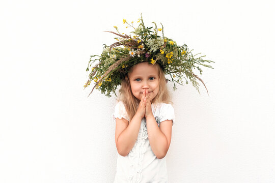 Little blonde girl in a wreath of wildflowers. Baby in white in a wreath on a white wall background